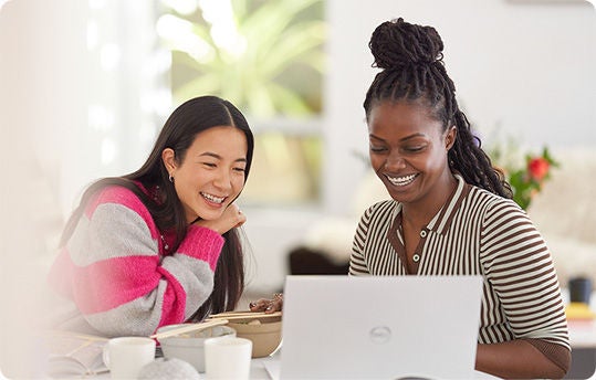 Two individuals are sitting at a table with a laptop and books. One person is wearing a striped shirt and has braided hair, while the other is wearing a sweater with pink and gray stripes.