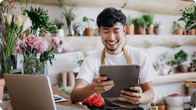 Person in a brown apron using a tablet at a table, surrounded by plants and flowers in a bright shop.