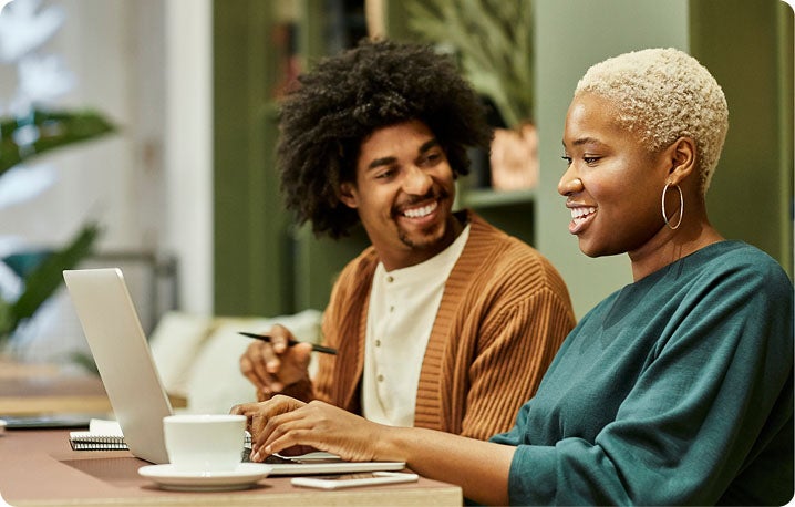Two people are sitting at a table with a laptop, notebook, and coffee cup. One person is holding a pen and appears to be discussing something with the other person.