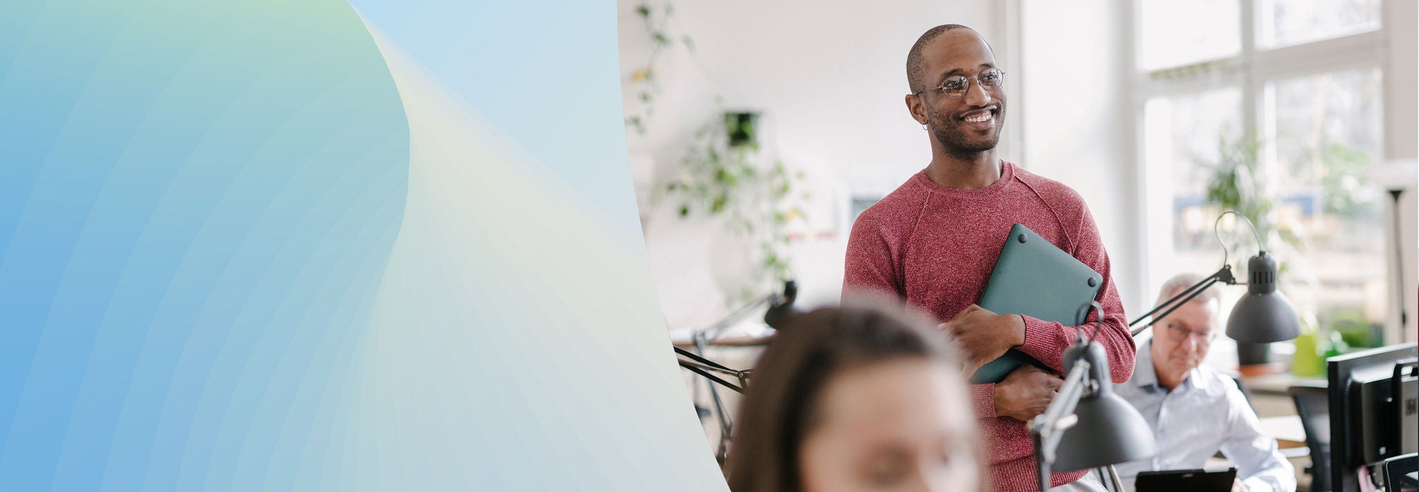 A man seated at a desk types on a keyboard while a woman stands beside him, leaning in to view the computer screen. Both are focused on the task, with another person working in the background.