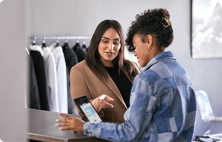 Two people are engaged in a conversation at a counter. One person, wearing a brown blazer, is gesturing with their hand while the other person, dressed in a blue checkered shirt, is holding a tablet.