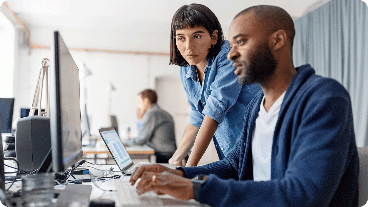 A man seated at a desk types on a keyboard while a woman stands beside him, leaning in to view the computer screen. Both are focused on the task, with another person working in the background.