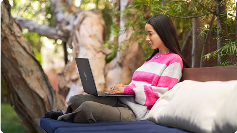 A person wearing a pink and white striped sweater is sitting on an outdoor bench with blue cushions, using a laptop.