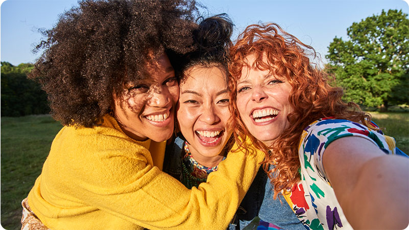 Three friends smile and embrace while taking a selfie outdoors on a sunny day, with green trees and grass in the background.